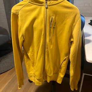 Gul hoodie från Sail Racing - Gul hoodie från Sail Racing med dragkedja framtill och praktiska fickor. Tröjan har en bröstficka med dragkedja och logga, samt huva och ribbade muddar. Perfekt för dig som gillar sportig och färgstark stil.