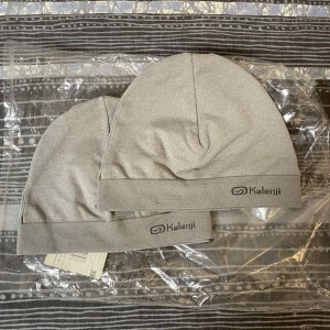 Grå mössa från Kalenji - Två stilrena grå mössor från Kalenji, perfekta för träning eller chill dagar. De har en enkel, rundad form med bred kant och diskret logga framtill. Materialet är mjukt och lätt, troligen polyester eller bomull, vilket gör dem sköna att bära.