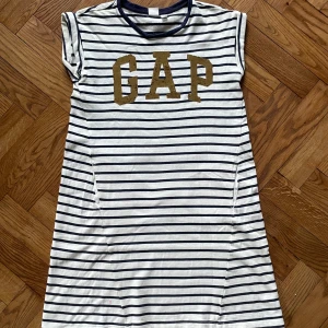 Randig kortklänning från GAP - Superchill vit och marinblå randig kortklänning från GAP med kontrastfärgade bokstäver framtill, ena varianten med guld och andra med röda paljetter. Mjuk bomull, rund hals och korta ärmar. Perfekt för dig som gillar enkel och sportig stil.