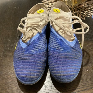 Nike Phantom blå fotbollsskor - Nike Phantom fotbollsskor i blått och vitt med gula detaljer och snörning. Ovandelen är stickad för bra passform och ventilation, och sulan har fasta dobbar för gräsplan. Snyggt randigt mönster på ovansidan och tydliga Nike-loggor på både sidor och häl.