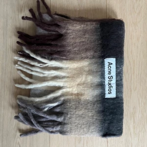 Mjuk halsduk Acne Studios i ull - Säljer en fluffig och lyxig halsduk från Acne Studios med breda färgblock i mörkbrun, beige och grå. Halsduken har långa fransar och stor Acne Studios-label. Perfekt för att ge din outfit en cool vibe under kalla dagar.