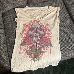 Vit topp/linne med döskalle och rosor - Säljer en vit topp från Miami ink med ett stort tryck framtill som visar en döskalle, röda rosor och coola detaljer i rosa och grönt. Toppen har v-ringning och är ärmlös, perfekt för dig som gillar edgy stil och statement-plagg.