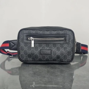 Svart axelväska från Gucci - Snygg svart axelväska från Gucci med klassiskt GG-mönster och silverfärgad dragkedja framtill. Väskan har en rektangulär form och justerbar rem i blått och rött. Materialet är en mix av skinn och syntet, perfekt för att bära dina essentials med stil.