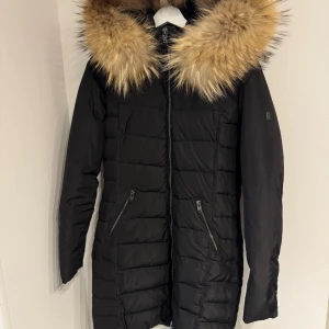 Svart dunjacka från Rockandblue - No sign of use. Svart lång dunjacka från Rock and Blue. Jackan har quiltad design, två snedställda fickor med dragkedja och hel dragkedja framtill. Perfekt för kalla vinterdagar och har en stilren look med diskret logga på ärmen.