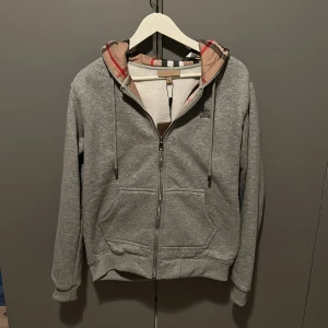 Grå Burberry hoodie med rutig huva - Grå hoodie från Burberry med klassisk rutig insida i beige, svart och röd. Tröjan har dragkedja, snörning vid huvan och broderad Burberry-logga på bröstet. Mjuk bomullskänsla och praktiska fickor framtill. Perfekt för dig som gillar stilren streetwear. Oanvänd 