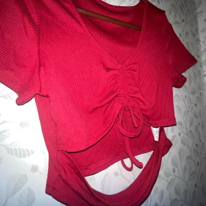 Röd croppad topp med snörning/Czerwony krótki top z koronkowymi detalami - Säljer en röd croppad topp med korta ärmar och snygg snörning framtill. Toppen har ribbad struktur, v-ringad hals och en cool öppen detalj nedtill. Perfekt för dig som vill sticka ut med en trendig look.