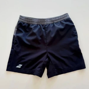 Svarta träningsshorts med grå kant - Snygga svarta träningsshorts med grå elastisk midja och en diskret logga på benet. Shortsen har en sportig look och är gjorda i lätt syntetmaterial som andas bra. Perfekta för gymmet eller löprundan, med bekväm passform och stilren design.