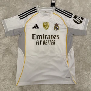 Real Madrid Mbappé 10 Adidas fotbollströja - Snygg vit Real Madrid fotbollströja från Adidas med Mbappé och nummer 10 på ryggen. Tröjan har svarta detaljer, gula linjer och grå partier på sidorna. Broderade klubbmärke och sponsorlogga framtill. Tillverkad i lätt och ventilerande material.