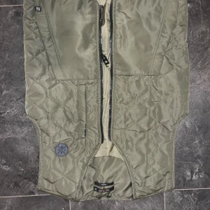 Olivgrön quiltad dunväst från Tretorn - Quiltad olivgrön dunväst från Tretorn med snyggt mönster och praktiska fickor. Västen har en rak passform, dragkedja framtill och en diskret logga på bröstet. Perfekt att slänga över en hoodie för extra värme under kyliga dagar.