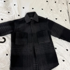 Svart rutig overshirt från H&M - Säljer en svart overshirt från H&M med diskret rutigt mönster. Jackan har två stora bröstfickor, klassisk krage och knäppning framtill. Materialet är mjukt och lite fluffigt, perfekt för lager-på-lager under hösten.