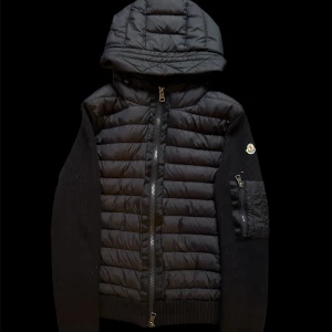 Moncler cardigan  - Svart pufferjacka från Moncler med stickade ärmar och quiltad front. Jackan har en stor huva, dragkedja framtill och en ficka med dragkedja på ärmen. Moncler-logga på vänster ärm. Perfekt för dig som vill ha en snygg och varm jacka med street vibe.