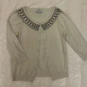 Beige kofta med pärlknappar och pärlbroderi - Supersöt beige kofta från Dear Princess med glittrande pärl- och paljettbroderi runt kragen. Koftan har pärlknappar framtill och trekvartsärmar. Har en liten lös pärla, se tredje bilden. 