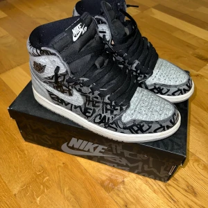 Nike Air Jordan 1 Retro High OG 41 - Nike Air Jordan 1 Retro High OG i svart, vit och grå med graffiti-inspirerat mönster och röda X på hälen. Skorna har högt skaft, snörning och rund tå. Snygg streetstil med unika detaljer och klassisk Air Jordan-logga på sidan. Kartongen till skorna ingår.