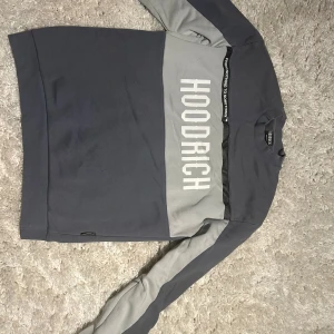 Grå crewneck från Hoodrich - Säljer en snygg grå och mörkgrå crewneck från Hoodrich med stort tryck på bröstet. Tröjan har lång ärm och klassisk passform, perfekt för dig som gillar streetwear. Materialet är mjukt och skönt, troligtvis bomull eller bomullsblandning.