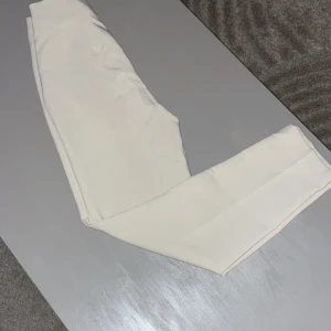 Vita kostymbyxor med pressveck - Snygga vita kostymbyxor med raka ben och pressveck framtill. Byxorna har ett slätt tyg med hög midja som ger en clean och stilren look. Perfekta för dig som vill ha en fräsch och modern stil. Aldrig använda! 