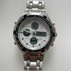 Silverfärgad kronografklocka EyeMonCroix - Behöver nytt batteri. Säljer en stilren kronografklocka från EyeMonCroix med silverfärgat metallarmband och vit urtavla. Klockan har digital display, dag- och datumvisning, alarm och tachymeter runt boetten. Perfekt för dig som gillar sportig design och många funktioner.