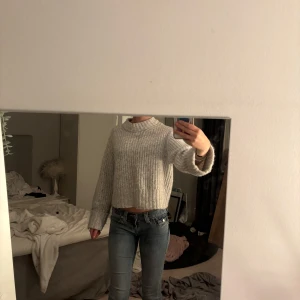 Ljusgrå croppad stickad tröja - Supermysig ljusgrå stickad tröja med croppad passform och breda ärmar. Tröjan har en högre krage och ribbade detaljer vid ärmslut och nederkant. Perfekt för lager-på-lager under kyliga dagar. Jätteskön tröja!