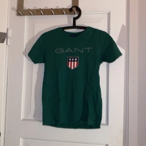 Grön GANT t-shirt med tryck - Snygg grön t-shirt från GANT med klassiskt tryck och sköld på bröstet. Modellen är kortärmad och har rund hals. Materialet är mjuk bomull som känns skönt mot huden. Perfekt för dig som gillar stilrena och sportiga plagg med en touch av preppy.
