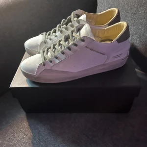 Vita sneakers från Crime London,  - Snygga vita sneakers från Crime London med grå detaljer och snörning. Skorna har en klassisk låg profil, rund tå och är tillverkade i skinn med beige insida. Perfekta för dig som gillar stilrena och moderna sneakers. Storlek 44 använda enbart en gång! 