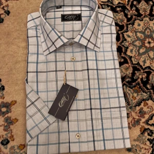 Rutig skjorta från Oscar of Sweden - Snygg rutig skjorta från Oscar of Sweden i ljusblått, vitt och grått. Skjortan är tillverkad i 100% bomull och har klassisk krage samt knäppning framtill. Perfekt för dig som gillar stilrena och fräscha mönster.