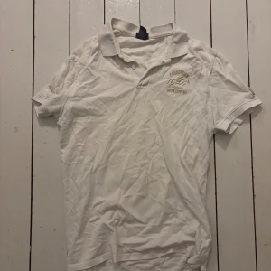 Vit polo t-shirt med broderi - Vit polo t-shirt med korta ärmar och broderad logga på bröstet. Klassisk krage med två knappar framtill. Perfekt för dig som gillar stilren och sportig look. Materialet känns som bomull och har en mjuk känsla.