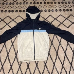 Blå och vit vindjacka från Lacoste - Snygg vindjacka från Lacoste i mörkblått och vitt med ljusblå detalj över bröstet och ryggen. Jackan har huva, dragkedja framtill och elastisk nederkant. Lacoste-logga på bröstet och tryck på ryggen. Perfekt för chill och streetwear.