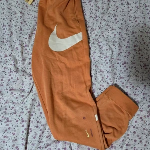 Nike mjukisbyxor orange - Säljer ett par orangea mjukisbyxor från Nike i storlek XL. Byxorna har stora vita Nike-loggor, snörning i midjan och ribbade muddar vid bensluten. Materialet är mjukt bomullstyg och byxorna har en loose passform. Perfekt för chill eller träning.