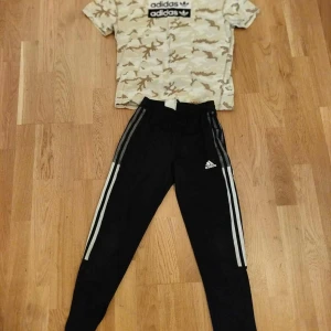 Svarta Adidas mjukisbyxor med ränder - Svarta mjukisbyxor från Adidas med klassiska vita ränder längs sidorna och Adidas-logga på låret. Byxorna har en slim passform och är perfekta för träning eller chill. Tillverkade i mjukt och bekvämt material.