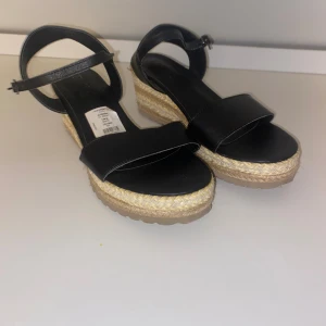 Svarta platåsandaler med flätad sula - Svarta sandaler med bred rem över foten och justerbart spänne vid ankeln. Skorna har en hög platåsula i flätad jute och gummisula undertill. Ovandelen är i syntetmaterial och sulan ger en somrig känsla. Perfekta för varma dagar. Lite sliten på bandet 