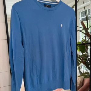 Blå tröja från Polo Ralph Lauren - Klassisk blå tröja från Polo Ralph Lauren med rund halsringning och diskret broderad logga i vitt på bröstet. Tröjan är långärmad och har ribbade muddar vid ärmslut och nederkant. Perfekt för en stilren och enkel look.