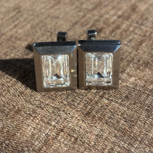 DK silvriga manschettknappar med kristall - Snygga rektangulära manschettknappar från DK i blankt rostfritt stål med en stor, glittrande kristall i mitten. Perfekt för att ge din skjorta en extra touch av stil. Märkta med 'STAINLESS STEEL' på baksidan och DK-logga på låset.