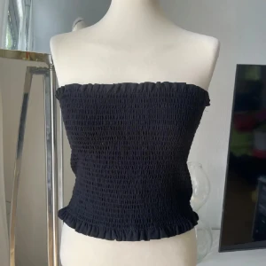 Svart smockad bandeau tubtopp - Säljer en svart bandeau tubtopp med smockad struktur och volangkant upptill och nedtill. Toppen är axelbandslös och har en tight passform som sitter snyggt mot kroppen. Perfekt för dig som gillar minimalistisk och stilren look.
