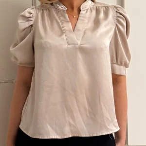 Champagnefärgad blus från Neo Noir - Säljer en stilren blus från Neo Noir i champagnefärgad satin med kort puffärm och v-ringad hals. Blusen har en mjuk glansig yta och lätt loose passform, perfekt för dig som gillar en chic och enkel look. Snygg att matcha med både jeans och kjol.