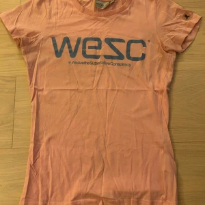 Ljusrosa WESC t-shirt med tryck - Snygg ljusrosa t-shirt från WESC i bomull med stort blått logotryck på bröstet. Klassisk rund hals och korta ärmar. Perfekt för dig som gillar streetwear och vill ha en enkel men cool look.