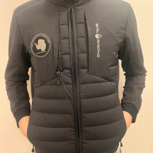 Svart pufferjacka från Sail Racing - Svart pufferjacka från Sail Racing med snygga logopatchar på bröstet och ärmen. Jackan har huva, två dragkedjor framtill och quiltad design. Materialet är polyester och jackan är långärmad med ribbade muddar. Perfekt för kalla dagar och har en clean, sportig look.