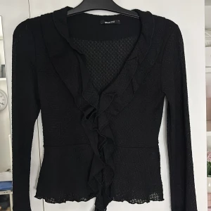 Svart volangblus Gina Tricot S - Supersnygg svart blus från Gina Tricot med volangdetaljer längs v-ringningen och framtill. Storlek S. Blusen har långärmade ärmar, lätt transparent material och vågig kant nedtill.