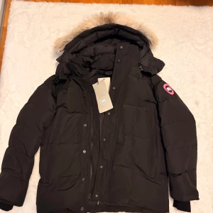Canada goose Wyndham  - Svart dunjacka från Canada Goose med fluffig beige päls på huvan och klassisk logga på ärmen. Jackan har dragkedja och knappar framtill, lång ärm och flera fickor. Perfekt för kalla vinterdagar med sin tjocka och värmande design.