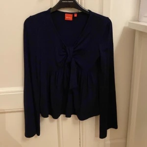 Mörkblå blus med knyt från RYVLS - Supersnygg mörkblå blus från RYVLS med lång ärm och v-ringning. Framtill finns ett coolt knyt som ger extra stilpoäng. Blusen har en lätt rynkad nederdel och är gjord i mjukt material som känns skönt mot huden. Perfekt för dig som gillar trendiga detaljer! Aldrig använd och säljer då den inte kommer till användning.