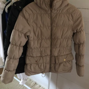 Beige pufferjacka med huva - Säljer en beige pufferjacka med huva och dragkedja framtill. Jackan har snörning i midjan, två stora fickor med knappar och quiltad design. Perfekt för kyliga dagar och har en snygg, enkel look som funkar till det mesta.