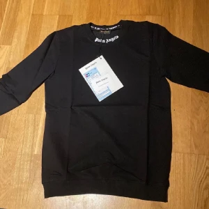 Svart Palm Angels sweatshirt - Svart sweatshirt från Palm Angels med stort vitt logotryck på ryggen och mindre logga vid halsen. Tröjan har rund halsringning och långa ärmar. Tillverkad i mjuk bomull för en skön och avslappnad stil. Perfekt för dig som gillar streetwear och vill sticka ut.