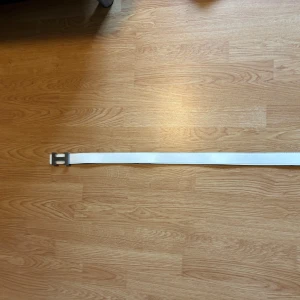 Vitt skärp med silverspänne - Snyggt vitt bälte med ett stort, stilrent silverspänne i form av ett H. Bältet är gjort i skinn och har en klassisk, rak form som passar till många olika outfits. Perfekt accessoar för att lyfta din look.