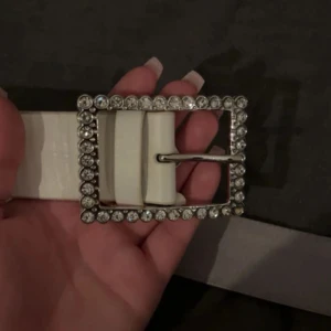 Vitt bälte med glittrigt spänne - Snyggt vitt bälte från Flash med ett stort, fyrkantigt spänne dekorerat med glittrande stenar. Bältet är brett och har en stilren look som verkligen sticker ut. Perfekt accessoar för att lyfta din outfit med lite extra bling.