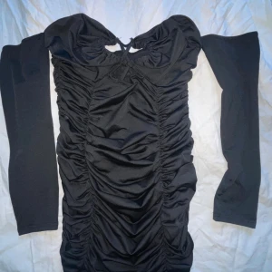 Svart offshoulder kortklänning - Snygg svart kortklänning med offshoulder och långa ärmar. Klänningen har en tight passform och är rynkad framtill för en cool och trendig look. Materialet känns mjukt och stretchigt, perfekt för att framhäva siluetten. Detalj med knyt upptill ger extra stilpoäng.