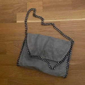 Grå axelväska med kedja och leopardfoder - Snygg grå axelväska i mocka med grov silverfärgad kedja som axelrem och runt kanterna. Insidan har ett coolt leopardmönstrat foder i brunt och beige samt en praktisk innerficka med dragkedja. Perfekt för dig som gillar detaljer och vill sticka ut.