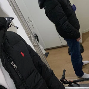 Svart pufferjacka från Polo Ralph Lauren - Svart pufferjacka från Polo Ralph Lauren med röd logga på bröstet. Jackan har dragkedja framtill, hög krage och justerbar snörning vid huvan. Perfekt för kalla dagar och har en snygg, modern look.