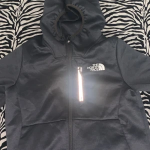 Grå kofta från The North Face - Grå kofta med huva från The North Face.Koftan har en snygg, stilren design med logga på bröstet och ryggen, hel dragkedja framtill samt en bröstficka med reflekterande dragkedja. Perfekt för dig som gillar streetwear och outdoor.