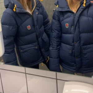 Mörkblå dunjacka från Fjällräven - Säljer en mörkblå dunjacka från Fjällräven med gul snörning i huvan och klassisk logga på bröstet. Jackan har stora fickor med knappar, dragkedja och huva. Perfekt för kalla dagar och riktigt snygg till jeans. Passar någon som vanligtvis bär S men Xs och M funkar!