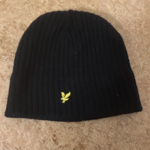 Svart ribbad mössa från Lyle & Scott - Svart ribbstickad mössa från Lyle & Scott med klassisk gul broderad logga framtill. Tillverkad i mjukt material och passar de flesta tack vare one size. Perfekt för kalla dagar när du vill hålla stilen enkel och snygg.