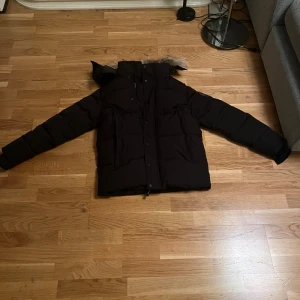 Svart dunjacka från Canada Goose - Säljer en svart dunjacka från Canada Goose med huva och snygg pälsdetalj. Jackan har långa ärmar, dragkedja och knappar framtill samt två stora fickor. Insidan är fodrad med blankt material och jackan är perfekt för kalla dagar.