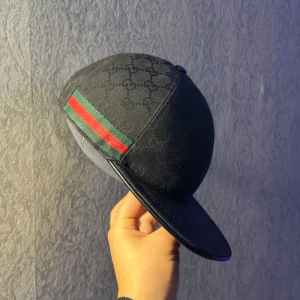 Svart Gucci keps med GG-mönster - Svart keps från Gucci med klassiskt GG-monogram över hela kepsen och en snygg grön-röd rand på sidan. Skärmen är böjd och kepsen har justerbart spänne bak. Materialet är bomull och polyester, perfekt för dig som vill ha en lyxig streetstil.